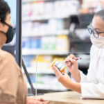 A female pharmacist of Hispanic descent is discussing medication with a customer from behind a plexiglass barrier during the coronavirus outbreak. They are both wearing a face mask to prevent the spread of germs.