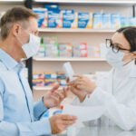 Male customer visitor giving medical prescription to female pharmacist in medical mask against coronavirus, while druggist advising new remedy pills antibiotics